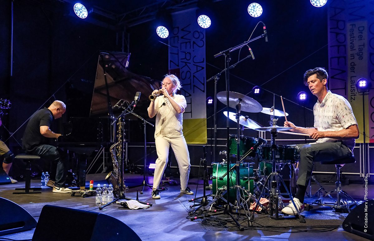 A vocalist in white with a pianist and a drummer on a blue-lit stage