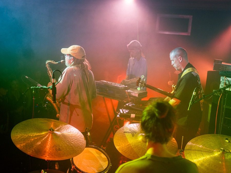 A band in warm stage light, a drummer in the foreground, saxophone and keys behind