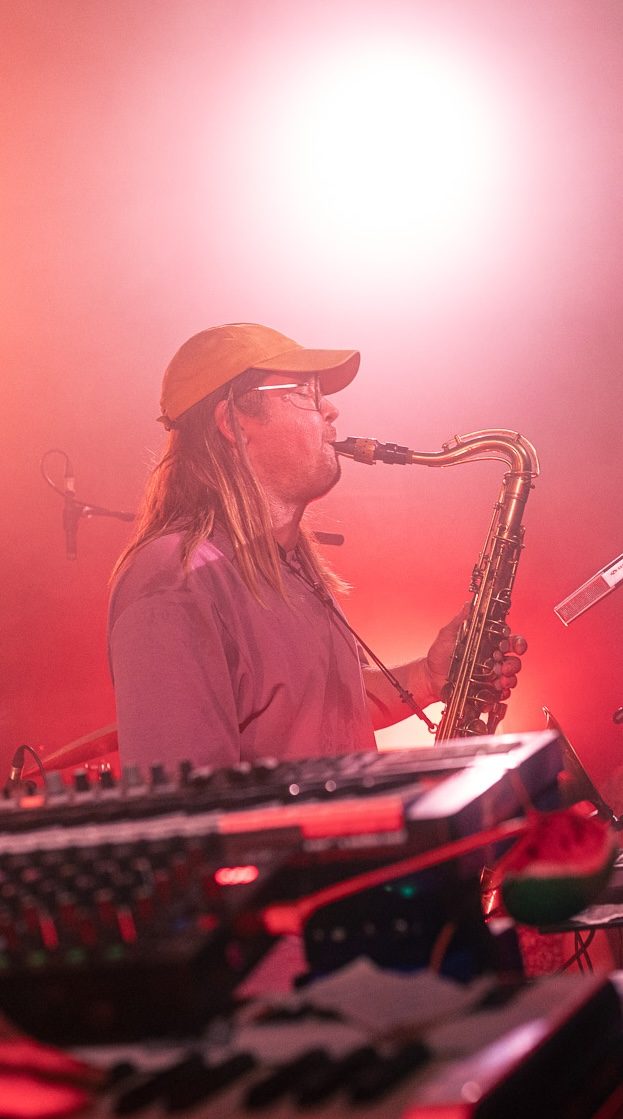 Saxophonist in a cap playing in red light behind a keyboard