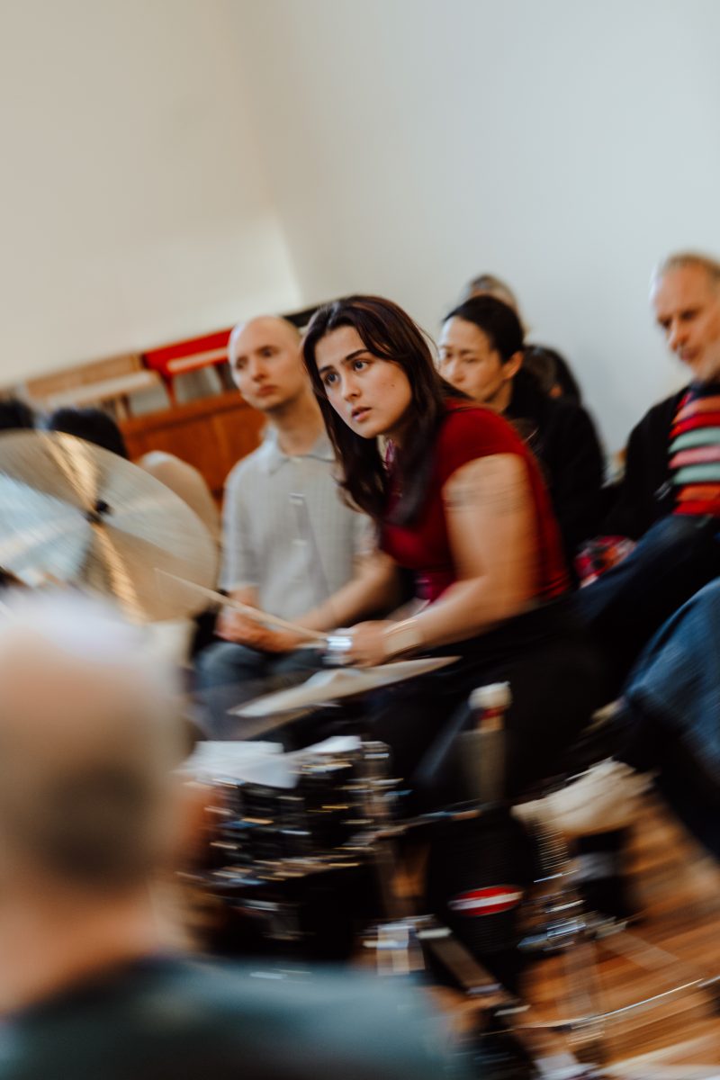 A drummer in a red top looks off to one side, with seated listeners close behind and a blurred cymbal in the foreground