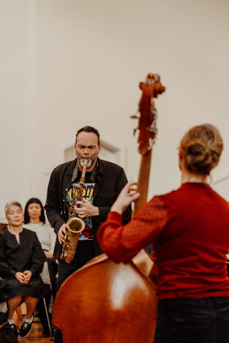 A saxophonist plays in front of a seated audience, with a double bass player seen from behind in the foreground