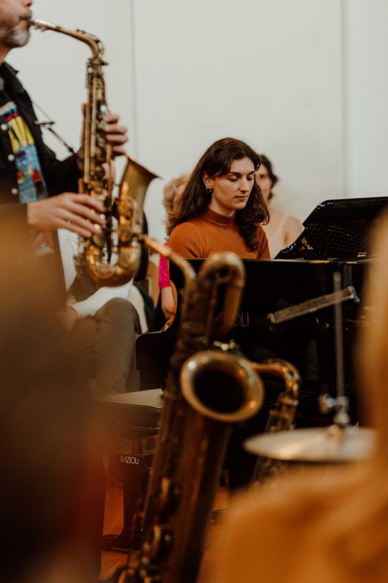 A pianist in a rust-coloured top looks down at the keys, framed by a saxophone and a baritone saxophone bell in the foreground