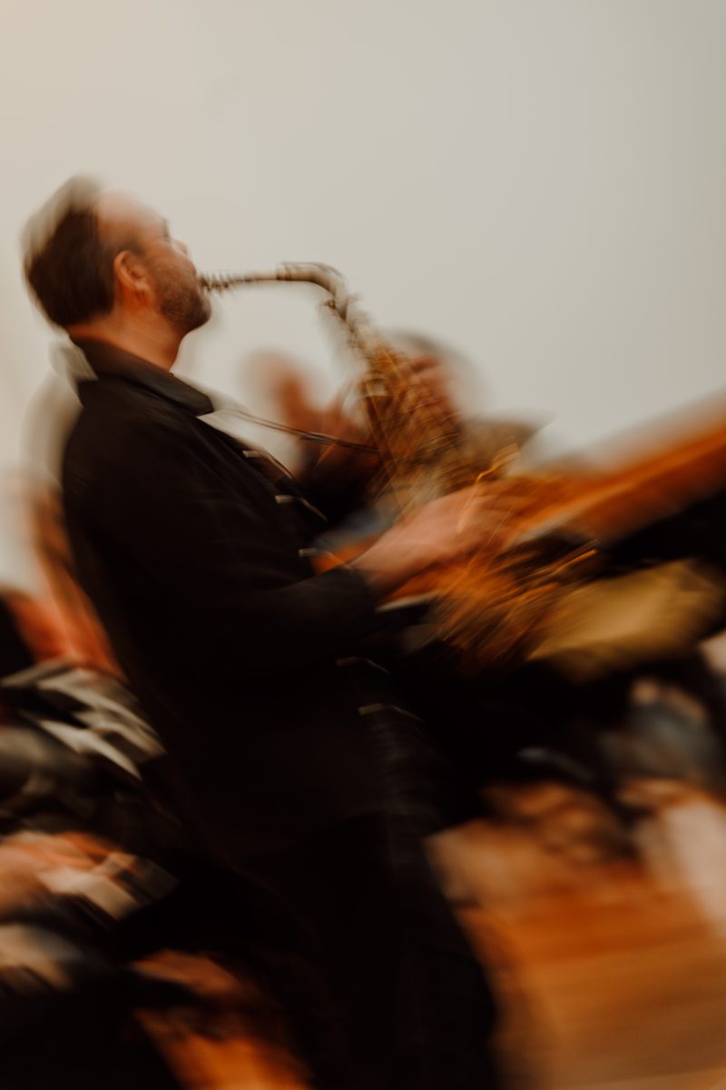 A saxophonist leaning back to play, smeared by motion blur against a pale wall