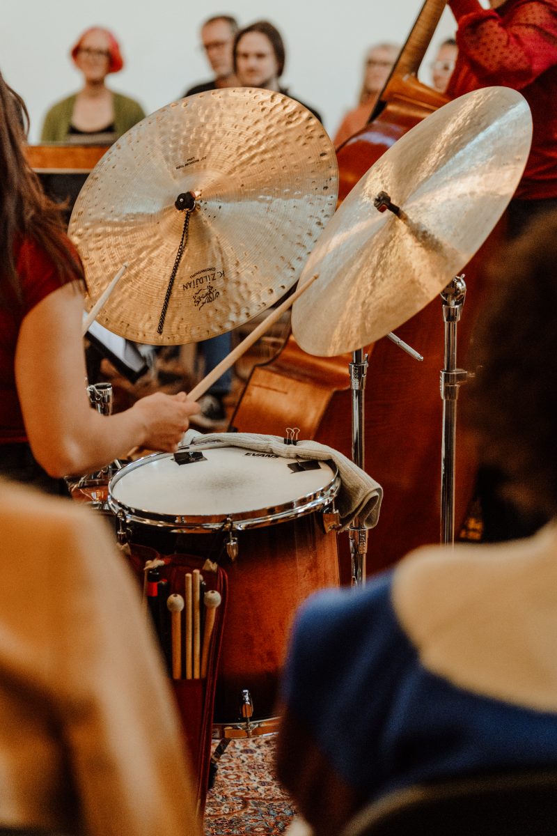 A drummer's hand and sticks over a snare drum and two cymbals, with a double bass and standing listeners behind