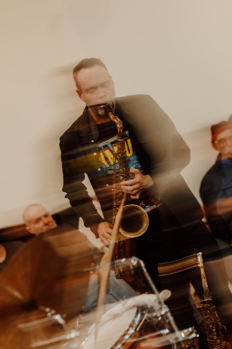 A saxophonist plays with eyes closed, streaked by motion blur, with a drum kit in the foreground and a listener to the right