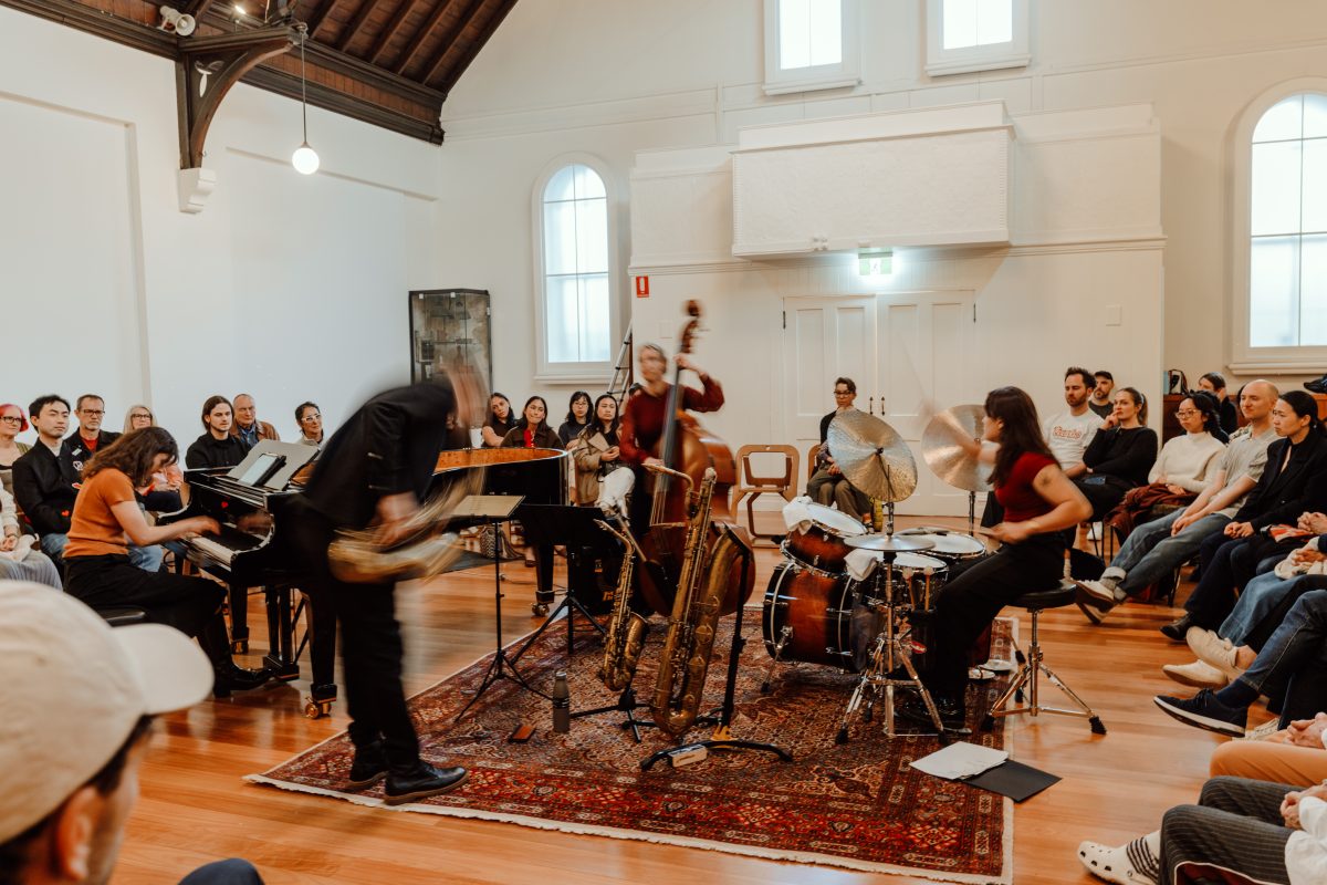 A wide view of a quartet on a patterned rug in a hall, the saxophonist bending forward in motion blur as the audience sits around them