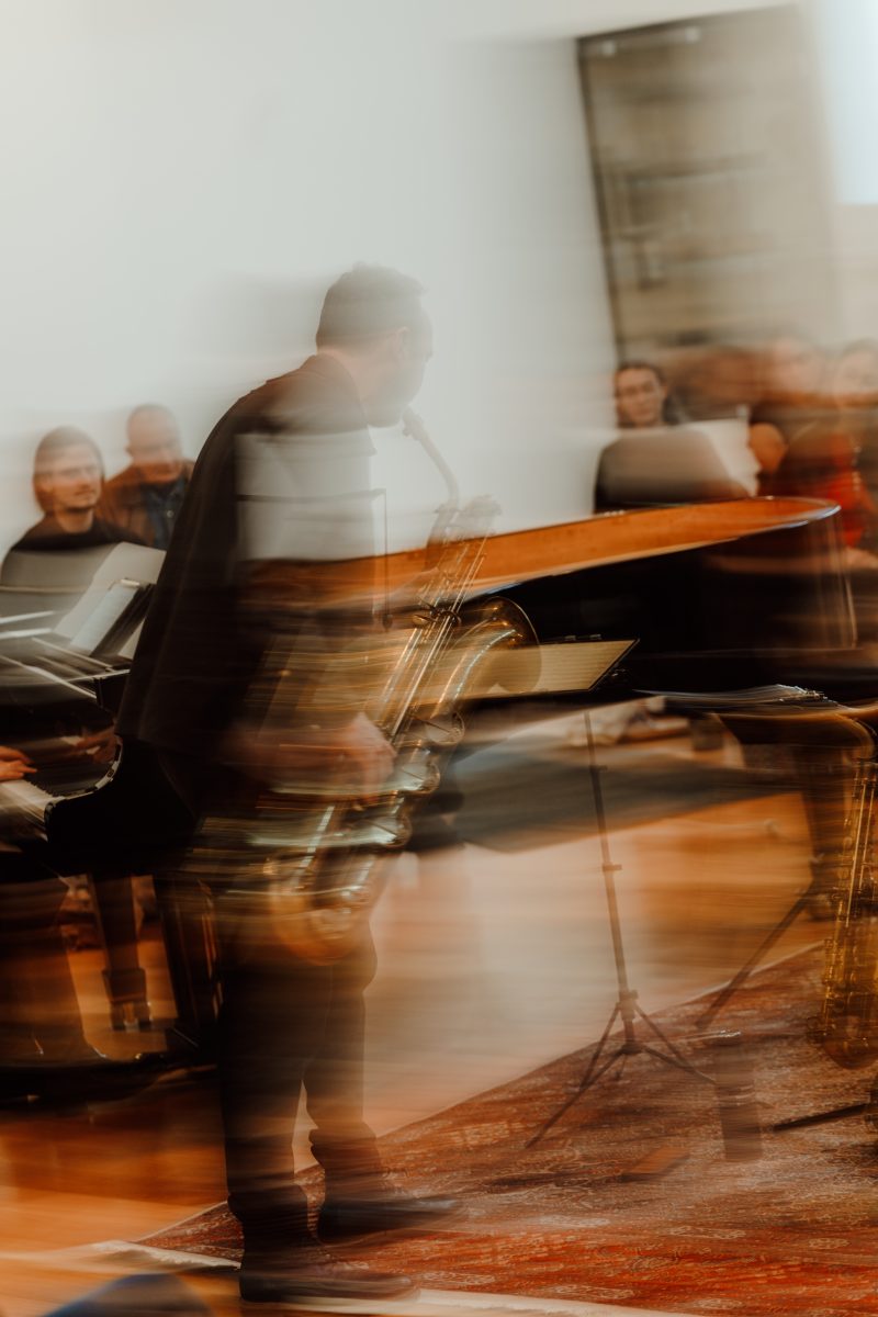 A saxophonist blurred by movement beside a grand piano, with listeners seated behind